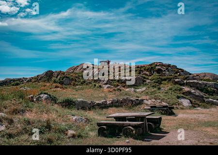 Un paesaggio panoramico con una collina rocciosa sormontata da una piccola struttura con tavoli da picnic in legno e panchine. Foto Stock