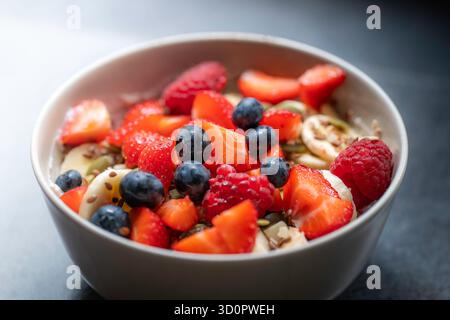 Farinata d'avena con frutta in una ciotola beige su un ripiano antracite. La ciotola è piena di fragole, mirtilli, lamponi, banane e semi di lino Foto Stock