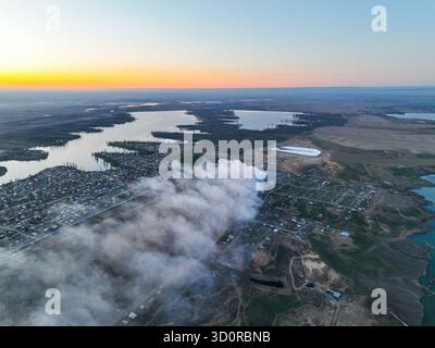Ampia ripresa aerea di un'area suburbana con fumo proveniente da un incendio che si diffonde sulle case e sui laghi vicini sotto un cielo limpido e colorato al tramonto Foto Stock