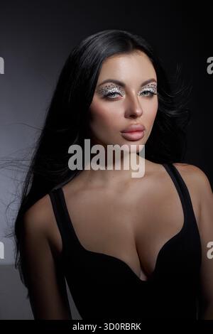 il modello si trova in uno studio scuro e presenta un trucco elegante con dettagli scintillanti. I suoi lunghi capelli scorrono con grazia, migliorando l'ar generale Foto Stock