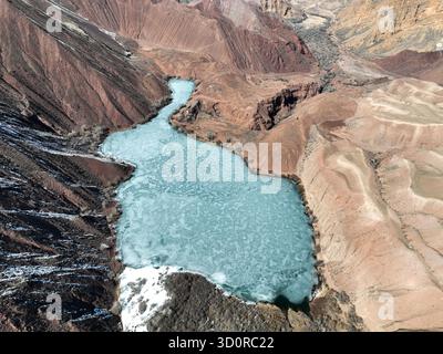 Alto scatto aereo verticale di un lago blu/turchese ghiacciato che si snoda attraverso una profonda valle delimitata da ripidi canyon erosi rossi e marroni a contrasto Foto Stock