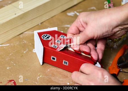 Una donna sta mettendo la neve decorativa su una piccola casa in legno dipinta di rosso e bianco su un tavolo marrone, lavorando con i dettagli, realizzando Foto Stock