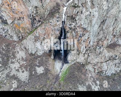 Vista aerea verticale diretta dall'alto, guardando verso il basso una cascata che cade giù da un'enorme parete rocciosa strutturata sul terreno roccioso sottostante Foto Stock