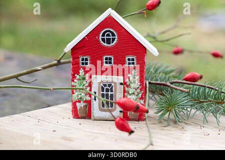 Affascinante casa in legno in miniatura, un elemento decorativo natalizio accoccolato tra le festose foglie invernali su una superficie rustica in legno. Foto Stock