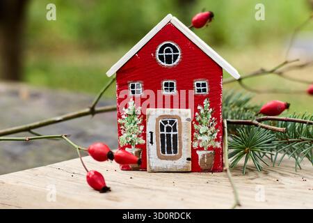 Affascinante casa rossa in miniatura con rifiniture bianche su una superficie in legno, circondata da rami con rosoni fianchi, che catturano una scena stravagante. Foto Stock