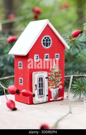 Incantevole arredamento natalizio: Una casa rossa in miniatura con tetto bianco e albero dipinto, adornata con rosa fianchi e rametti Evergreen. Foto Stock