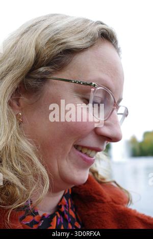 Una donna dai capelli biondi ondulati che indossa gli occhiali viene mostrata in primo piano. Sta sorridendo e guardando verso il basso. È vestita con una top a colorata Foto Stock