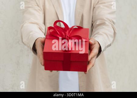 Uomo che regala una confezione regalo rossa legata al nastro e indossa una camicia beige per il compleanno o San Valentino. Il regalo suggerisce una sorpresa o un'occasione speciale. Foto Stock