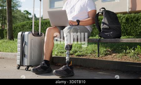 Una persona con una gamba protesica siede su una panchina utilizzando un notebook. Una valigia è accanto a loro in un parco. Foto Stock