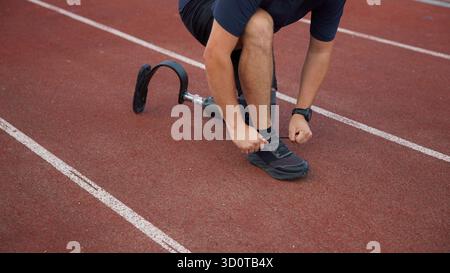 Un uomo si inginocchia su una pista da corsa, regolando il pizzo della sua scarpa mentre usa una gamba protesica, preparandosi a correre. Foto Stock