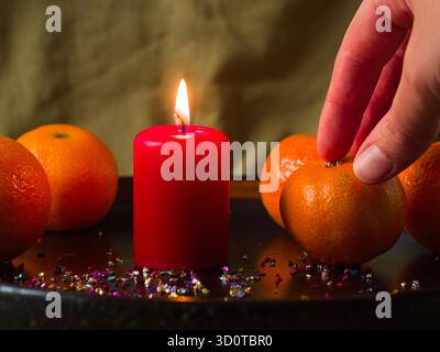 La mano di una persona posiziona un mandarino fresco accanto a una candela rossa accesa su un vassoio festivo, preparando un intimo Natale o la decorazione della tavola di Capodanno Foto Stock