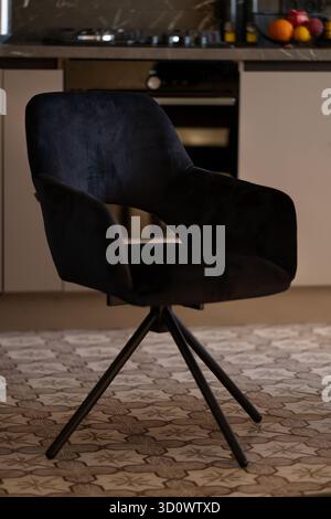 Un'elegante sedia da pranzo in velluto blu scuro con base in metallo nero si trova in una cucina ben illuminata, caratterizzata da armadi bianchi e un tappeto a motivi geometrici. Foto Stock