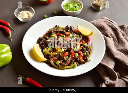 Mescolare il controfiletto di manzo fritto a fette sottili con le verdure su un piatto bianco su fondo marrone scuro. Vista ravvicinata Foto Stock