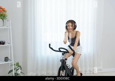 La giovane donna felice ama l'attività fisica a casa per le persone, cavalcare la cyclette con le cuffie, sorridere ampiamente mentre ascolti la musica, abbracciando la Foto Stock