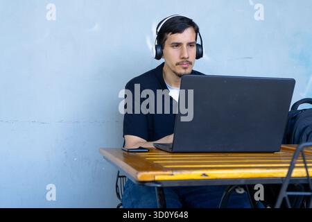 Uomo che si concentra sul proprio laptop, indossa le cuffie e lavora a distanza a un tavolo. Rappresenta uno stile di vita nomade digitale, uno studente o un freelance Foto Stock