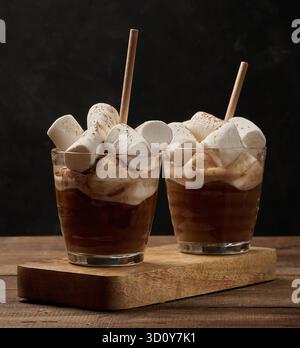 Due bicchieri di cioccolata calda con soffici marshmallow e una cannuccia su un vassoio di legno Un accogliente drink invernale su uno sfondo scuro. Foto Stock