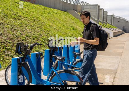 Giovane uomo che indossa uno zaino che interagisce con una stazione di condivisione delle biciclette utilizzando un'app mobile in una giornata di sole Foto Stock