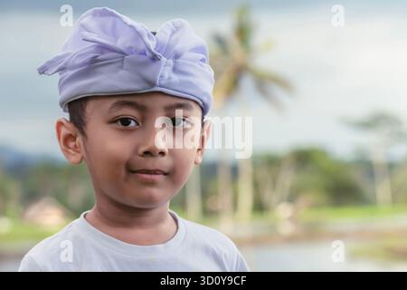 Ritratto di un bambino balinese con un sorriso fresco e sottile in abiti cerimoniali bianchi e un campo di riso leggermente sfocato sullo sfondo il giorno delle Galungan. Foto Stock