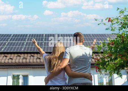 Giovane coppia che abbraccia, guardando i pannelli solari sul tetto della loro casa. Foto Stock
