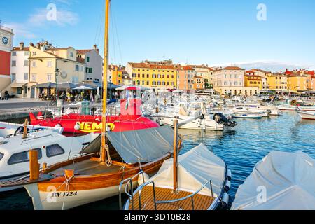 Negozi e caffetterie lungo la piazza principale di Trg Marsala presso il colorato villaggio di Rovigno, in Croazia, con barche nel porto. Foto Stock
