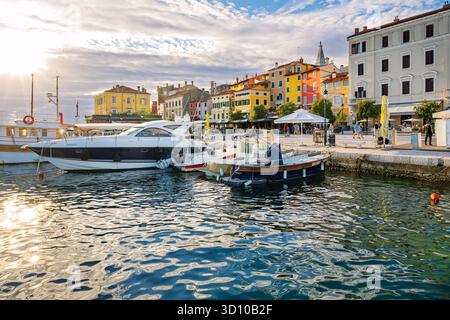 Barche ancorate lungo il lungomare con edifici colorati pieni di caffetterie e negozi sul marciapiede nella zona Trg Marsala Tita di Rovigno, Croazia Foto Stock
