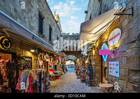 Negozi di souvenir, boutique e caffetterie fiancheggiano i vicoli stretti con la porta medievale di Arnoldo e la vista sul mare, all'interno del castello fortificato di Rodi, Grecia. Foto Stock