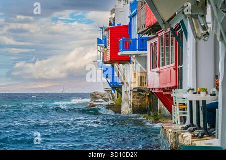 Edifici colorati con balconi si affacciano sul Mar Egeo accanto a caffetterie all'aperto mentre le onde colpiscono la costa nel quartiere Little Venice di Mykonos, in Grecia. Foto Stock