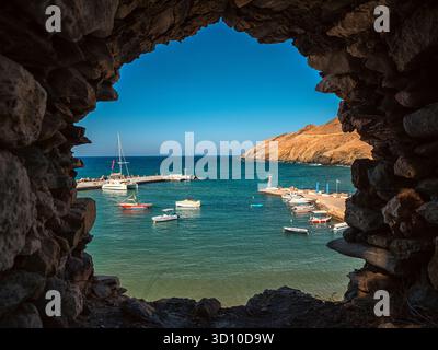 Panormos, Grecia - 9 settembre 2025: Vista panoramica del piccolo porto e delle barche viste attraverso l'antica finestra a muro in pietra nel villaggio Panormos sull'isola di Creta Foto Stock