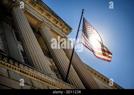 Bandiera DEGLI STATI UNITI retroilluminata dal sole sull'Herbert C Hoover Federal Building Washington DC // WASHINGTON DC - la bandiera degli Stati Uniti è retroilluminata dal sole sull'Herbert C. Hoover Federal Building, sede del Dipartimento del commercio degli Stati Uniti. Situata in Pennsylvania Avenue NW a Washington DC, questa struttura monumentale è una componente chiave del Triangolo Federale. Progettato in stile neoclassico dall'architetto Waddy B. Wood, l'edificio è stato costruito tra il 1928 e il 1932. La sua grande facciata e la sua posizione prominente riflettono il suo ruolo nella capitale della nazione. Foto Stock