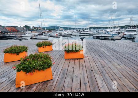 Oslo, Norvegia - 8 settembre 2025: Vasi di fiori di colore arancione su argini e barche ormeggiate nel porto del distretto di Aker Brygge Foto Stock