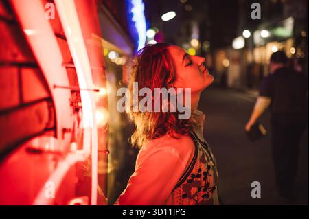 Giovane donna appoggiata contro il cartello al neon di notte in Urban Street Foto Stock