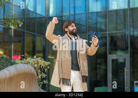 Un uomo d'affari indiano entusiasta e stupito che utilizza un dispositivo di telefonia mobile un uomo online scioccato in centro città vince una scommessa su smartphone festeggia il successo della vittoria. Un tizio in piedi sulla strada urbana del centro Foto Stock