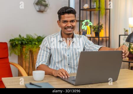L'uomo indiano a casa apre il portatile inizia le attività prende un sorso di caffè e inizia un lavoro concentrato. Il ragazzo arabo alla scrivania controlla i tipi di elenchi di progetti sulla tastiera mantiene un ritmo costante mostra i progressi e l'umore molto buono Foto Stock