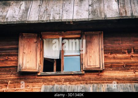 Finestra con persiane aperte e semplici tende bianche su una facciata in legno con capanna in legno, che mostra un'architettura rustica Foto Stock