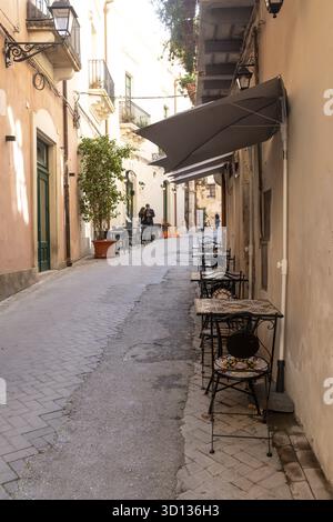 Syrakus, Sizilien - Italia - 04-08-2025: Tavoli da caffe' vuoti in ferro battuto con cime colorate a mosaico fiancheggiano una stretta e soleggiata strada a Siracusa, circondata Foto Stock
