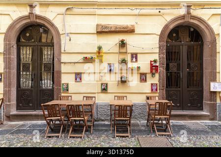 Bosa - Sardinien - Italien - 02-28-2025: Veduta di una colorata facciata bistro con tavoli e sedie in legno, porte incorniciate, arte murale, piante in vaso, piccole Foto Stock