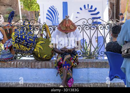 Chefchaouen, Chefchaouen - Marocco - 08-20-2024, uomo in abiti tradizionali con un cappello di paglia decorato siede su un muro di pietra, guardando il suo telefono. Col Foto Stock