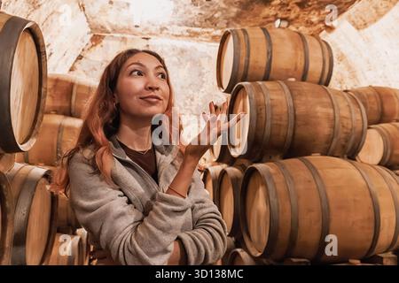 Woman si erge tra botti di rovere impilate in una cantina tradizionale, esplorando la produzione di vino e degustando il turismo Foto Stock
