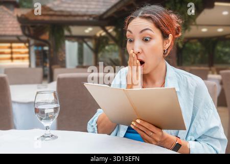 Donna che sembrava sorpresa e scioccata mentre teneva un menu, si rendeva conto dei prezzi costosi in un ristorante all'aperto Foto Stock