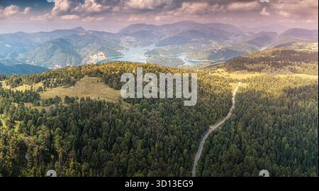 Vista panoramica aerea di un paesaggio montano caratterizzato da un vasto lago, foreste verdi e colline ondulate con una strada tortuosa Foto Stock