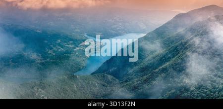 Vista aerea panoramica che mostra le montagne e un lago a valle circondato da una lussureggiante foresta verde, con nuvole e nebbia Foto Stock