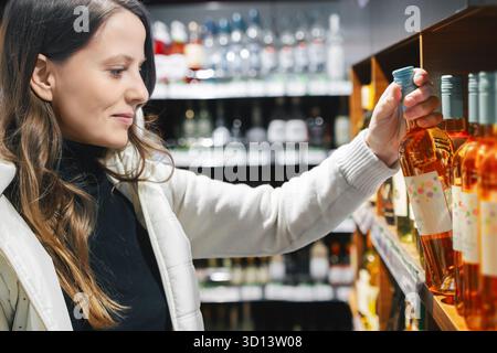 Una donna allegra e bella ama fare shopping mentre preleva una bottiglia di vino rosa dal bancone. L'ambiente è un negozio ben rifornito con un caldo Foto Stock