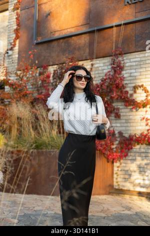 Elegante signora con capelli ricci e occhiali da sole in piedi all'aperto in autunno, vestita con un'accogliente moda di stagione. Foto Stock