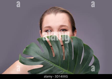 Donna adulta sorridente caucasica che sbircia dalla foglia di monstera su sfondo grigio. || modello rilasciato Foto Stock