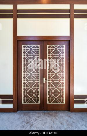 Doppia porta in legno decorato con motivi geometrici islamici dorati e maniglia in ottone, incastonata in una parete di colore chiaro con finiture in legno scuro, sopra un tappeto grigio Foto Stock