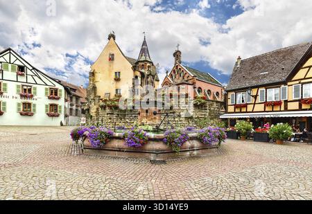 Eguisheim, Francia - 8 luglio 2019: Piazza con colorate case tradizionali francesi e fontana con statua di papa Leone IX a Eguisheim a Eguisheim TOW Foto Stock