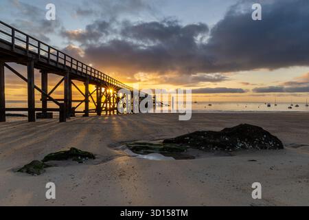 Un molo di legno si estende su una tranquilla spiaggia all'alba, gettando lunghe ombre sulla sabbia. Le barche galleggiano sull'acqua calma mentre le nuvole spettacolari brillano di golo Foto Stock