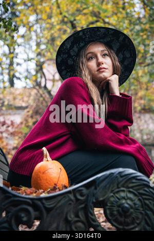 Una giovane donna con un maglione di Borgogna e un cappello nero siede su una panchina del parco accanto a una piccola zucca, circondata da fogliame autunnale Foto Stock