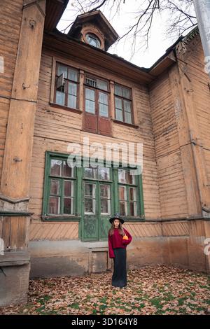 Una donna con un cappello da strega e un maglione Borgogna tiene una zucca di fronte a una vecchia casa di legno con finestre verdi Foto Stock