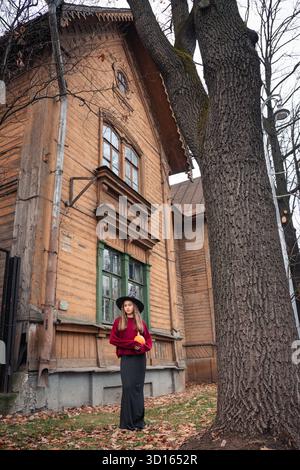Una donna con un cappello da strega e un maglione Borgogna tiene una zucca di fronte a una vecchia casa di legno con finestre verdi Foto Stock